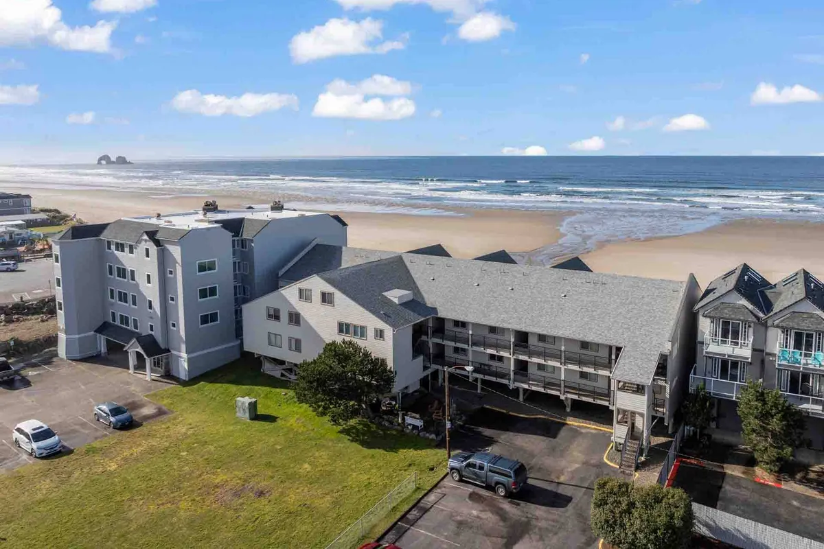 A wide exterior shot featuring the parking area in front, the beachfront, and the surrounding buildings, all set against a tranquil backdrop. The image captures the sense of arrival and the easy transition from parking to the unit's entrance.