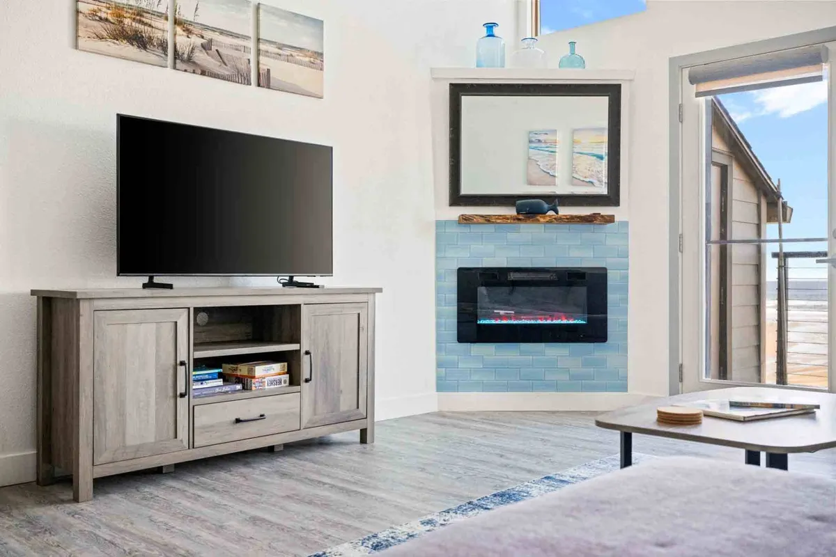 A cozy, sunlit living area featuring a comfortable seating arrangement around a modern fireplace, a flat-screen TV, and a large window that lets in abundant natural light. The coffee table sits at the center, inviting quiet moments of reading or creative work while enjoying a view of the outdoors.