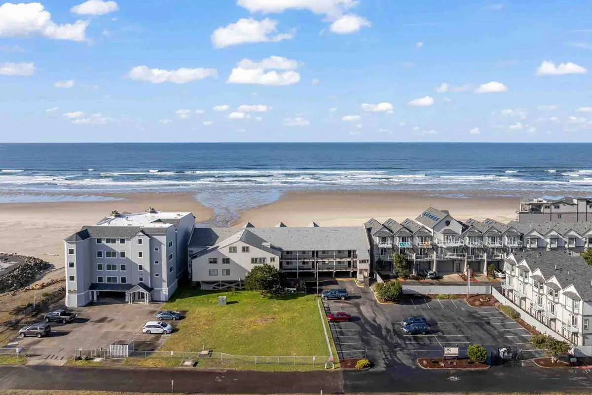 A panoramic exterior view captures the beach just steps from nearby buildings, with gentle waves lapping at the sand and the golden glow of sunset beginning to appear. The scene feels both tranquil and accessible, highlighting the close proximity between the property and the shoreline.