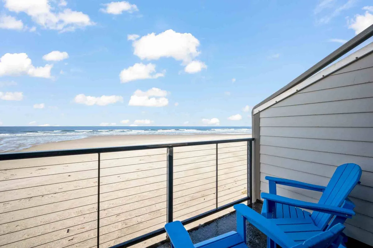 Two blue Adirondack chairs face out over a sweeping ocean view, with soft clouds in the sky and the expanse of the Pacific stretching to the horizon. The chairs are placed on a deck or balcony, inviting guests to relax and soak in the beauty of the setting sun or enjoy a peaceful morning.