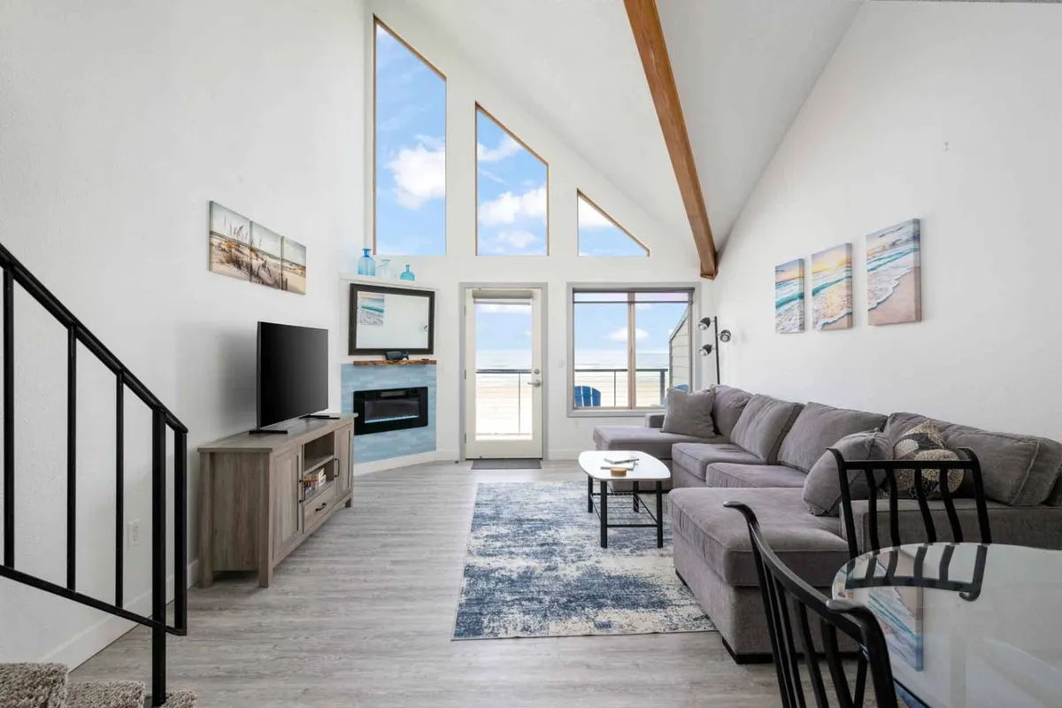 The image showcases a cozy, light-filled living area with a large sectional sofa facing a sleek modern fireplace. Expansive windows flood the room with natural light and frame sweeping views of the ocean, while tasteful artwork and inviting décor create a warm, welcoming atmosphere ideal for relaxation and socializing.