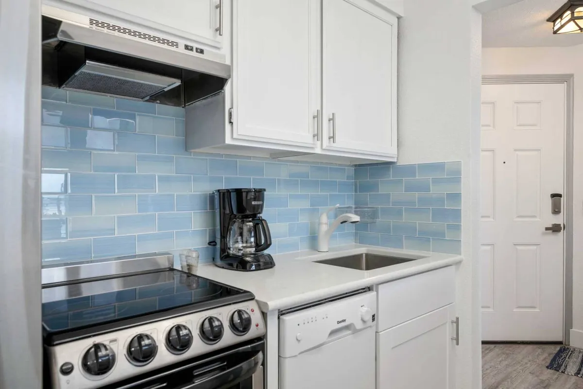 A bright, modern kitchen featuring a stainless steel stove, blue tiled backsplash, coffee maker, white cabinets, and a sink. The setup is inviting for making coffee, preparing meals, or gathering with friends to chat and enjoy breakfast before heading out to explore town.