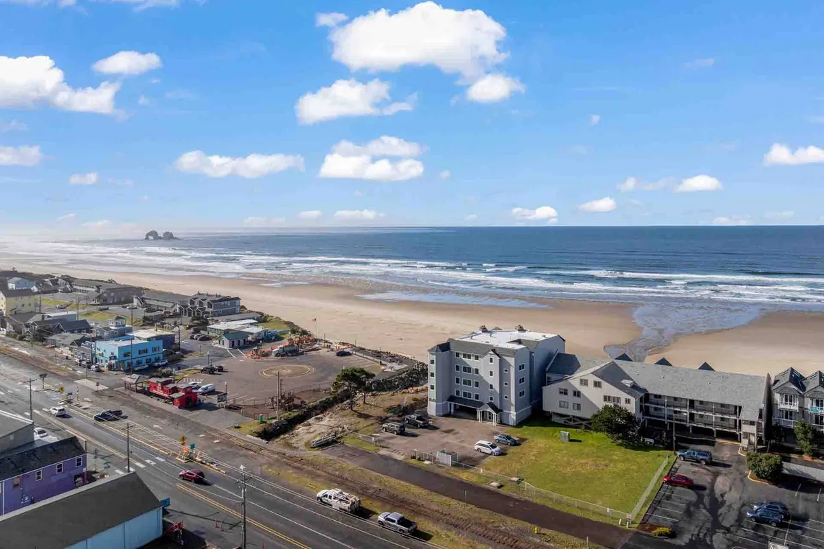 This exterior image captures a beautiful stretch of sandy beach with ocean waves, nearby buildings, and a road. The view suggests easy beach access and the presence of vacation-friendly infrastructure, perfect for families heading straight out for classic beach activities.