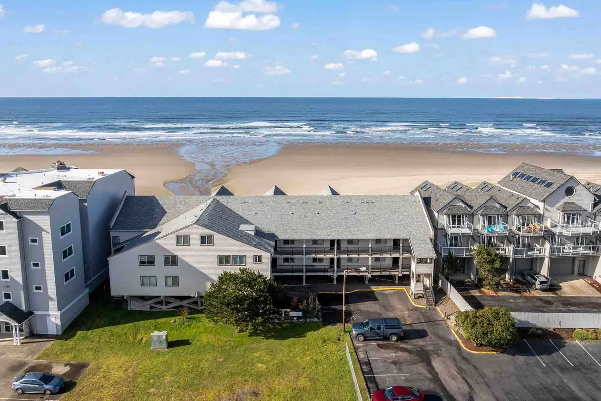 This exterior photo captures the building's architecture with the adjacent parking area clearly visible in the foreground. The beach and ocean extend beyond, providing context for proximity to both the parking lot and the coastal environment.