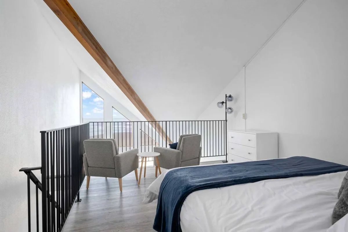 A serene loft bedroom featuring a comfortable bed with a blue throw, two armchairs with a small table, and large windows that flood the space with natural light. Exposed wooden beams add warmth and character, while the windows offer glimpses of the ocean beyond.