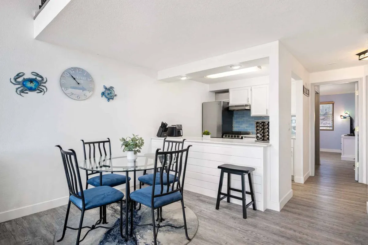 A stylish dining area with a glass dining table surrounded by blue chairs, accented with decorative wall art and glimpses of the kitchen and appliances nearby. The space is bathed in natural light, creating a welcoming environment for meals and gatherings.