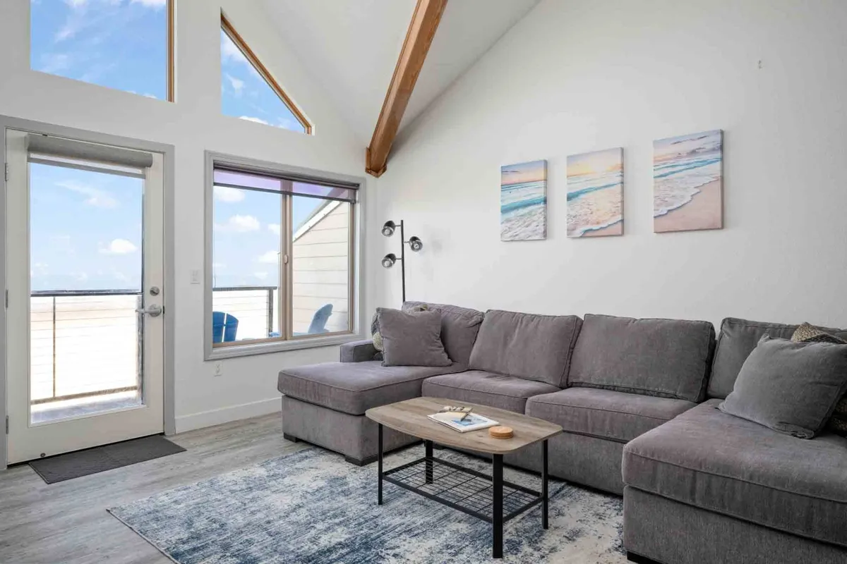 A cozy sectional sofa in an open living area, accented by wall art and a window that frames an inviting view. The space is styled for comfort and group lounging, perfect for board games or watching the sunset together.